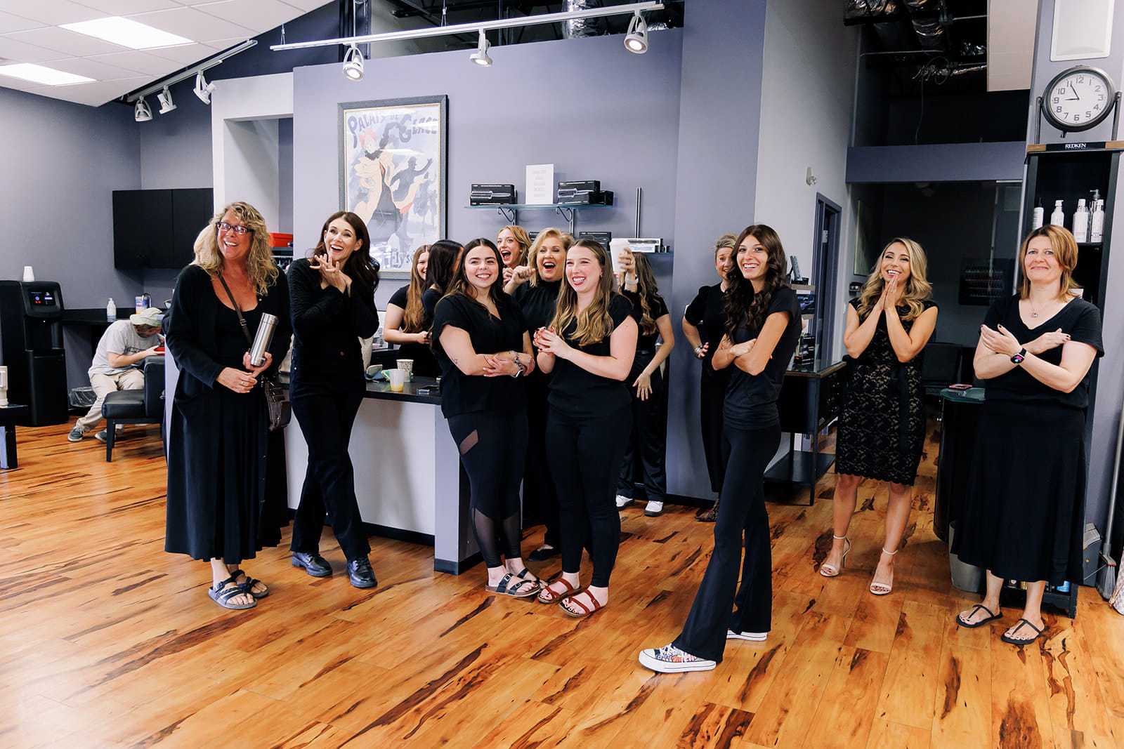 Group of smiling women in a salon, dressed in black, posing together on a wooden floor.